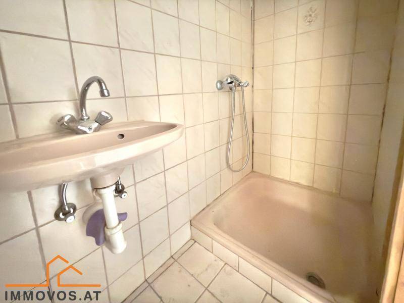 Kleines Badezimmer mit beigen Fliesen, Waschbecken und einer Duschwanne, die renoviert werden muss.