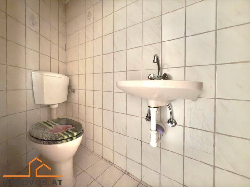Einfaches WC mit weißen Fliesen, Toilette und Waschbecken, das eine Modernisierung benötigt.