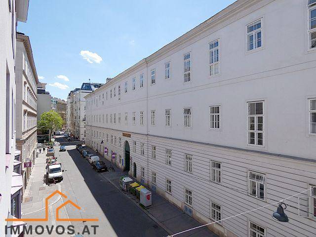 Lange, weiße Gebäudefassade mit vielen Fenstern entlang einer ruhigen Stadtstraße mit parkenden Autos.