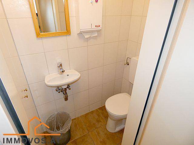 Gästetoilette mit kleinem Waschbecken, WC und braunem Fliesenboden, umgeben von weißen Wandfliesen.