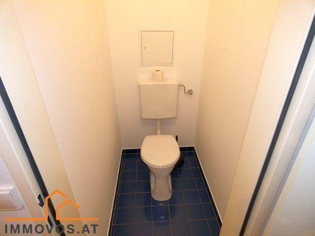 Separate Toilette mit WC und blauen Fliesen auf dem Boden, umgeben von schlichten weißen Wänden.