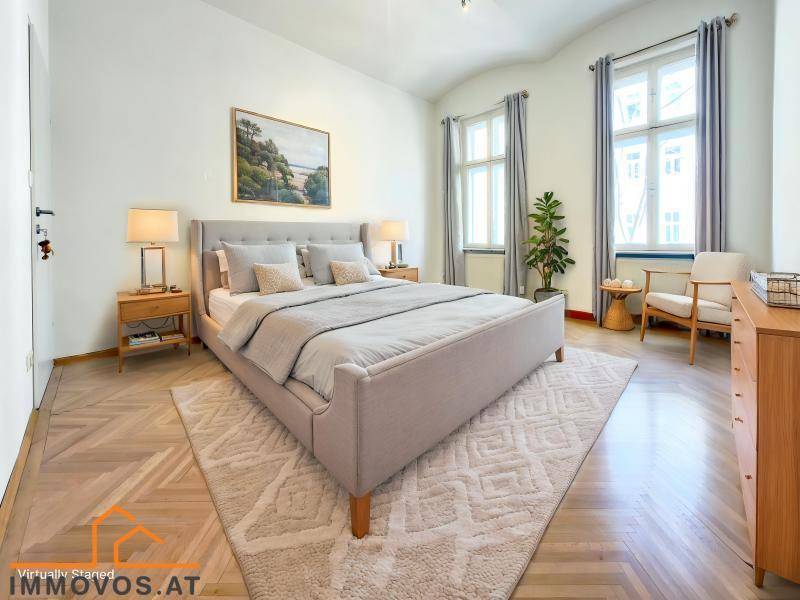 Elegantes Schlafzimmer mit Fischgrätparkett, bequemen Doppelbett und zwei hohen Fenstern für eine helle Atmosphäre.