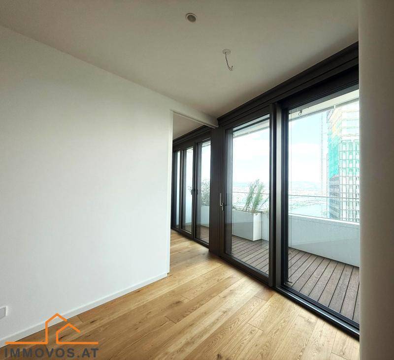 Lichtdurchfluteter Raum mit Holzboden und raumhohen Fenstern, die einen weiten Blick und Zugang zum Balkon ermöglichen.