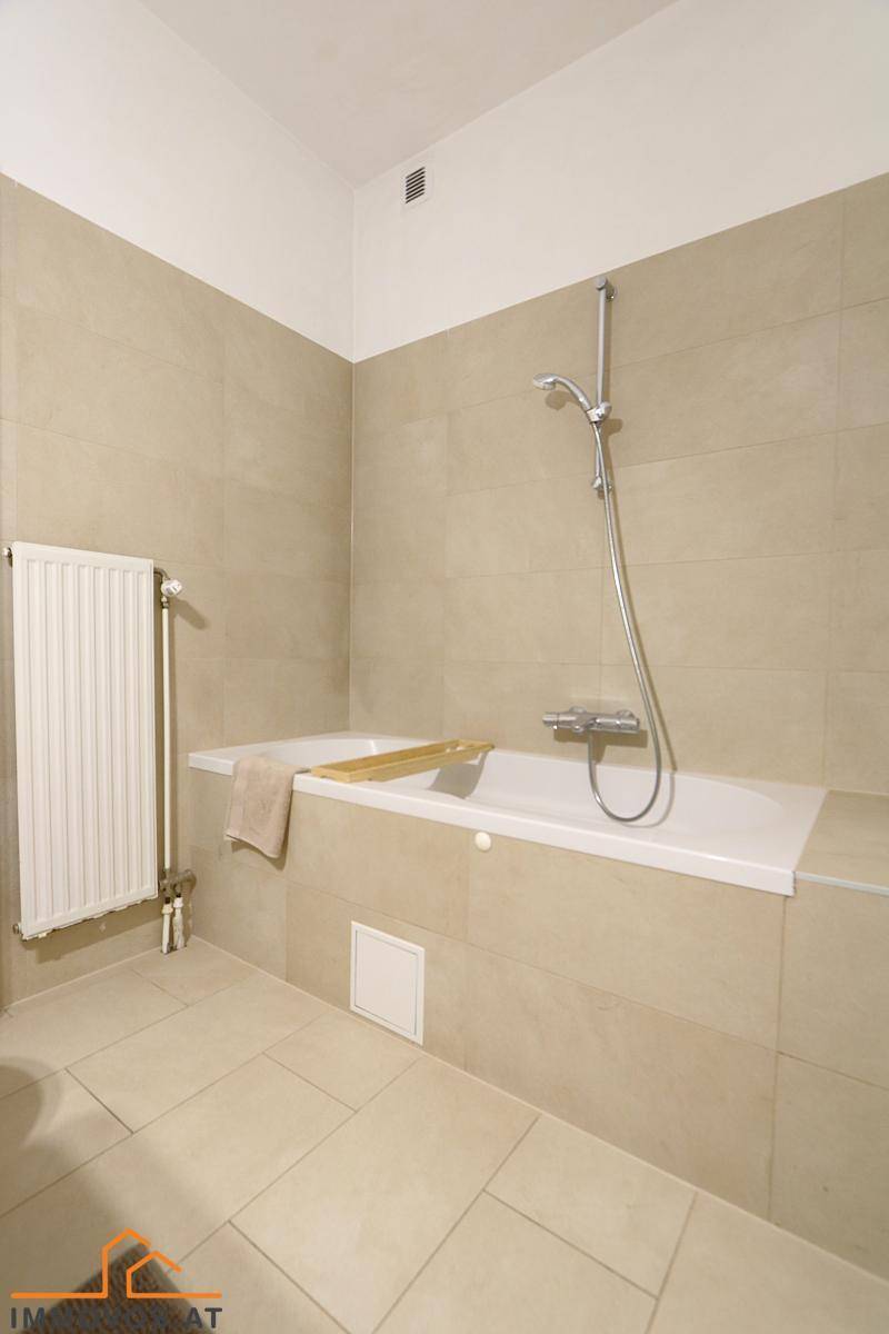 Badezimmer mit Badewanne, Duscharmatur und beigefarbenen Wand- und Bodenfliesen.