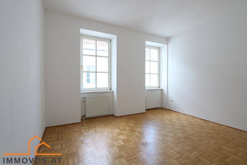 Helles, leeres Schlafzimmer mit Fischgrätparkettboden und zwei Fenstern.