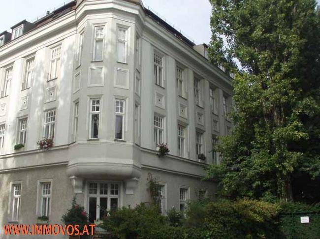Historisches Eckgebäude mit vielen Fenstern, umgeben von üppigem Grün und Bäumen.