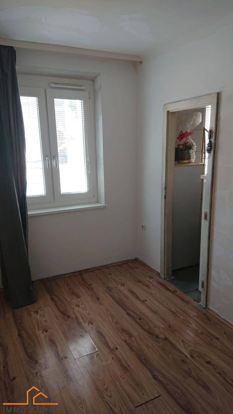 Leerer Raum mit Holzboden, einem Fenster mit Vorhang und Zugang zu einem weiteren Zimmer.