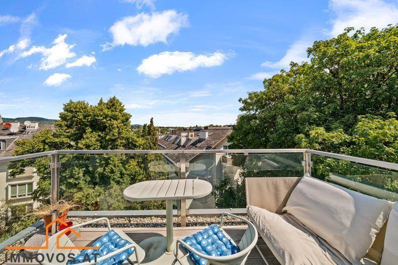 Sonniger Balkon mit Glasgeländer, Sitzgelegenheiten und weitem Blick über die Dächer und Bäume.