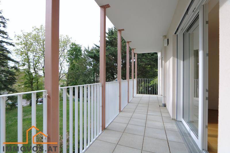 Langer Balkon mit Fliesenboden und weißem Geländer, umgeben von grünen Bäumen.