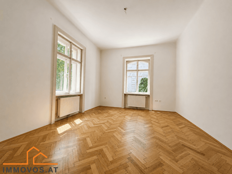 Helles, leeres Zimmer mit Fischgrätparkett und zwei großen Fenstern, ideal als Schlaf- oder Arbeitszimmer.