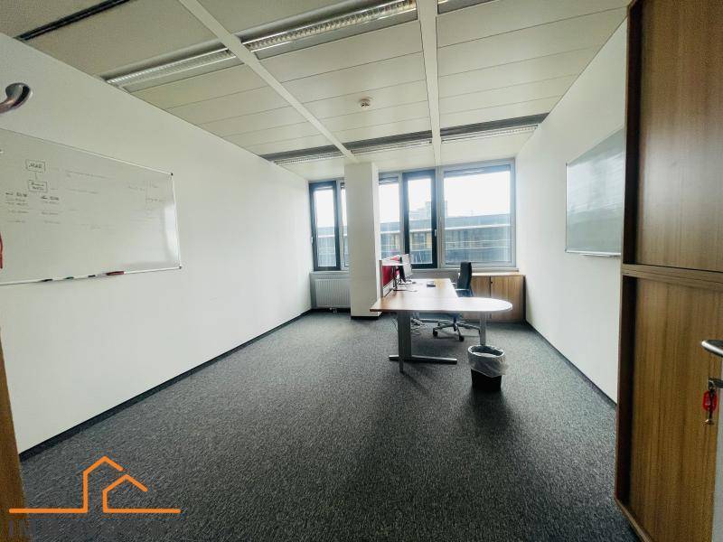 Leerer Büroraum mit Teppichboden, großen Fenstern und einem einzelnen Schreibtisch in der Ecke.