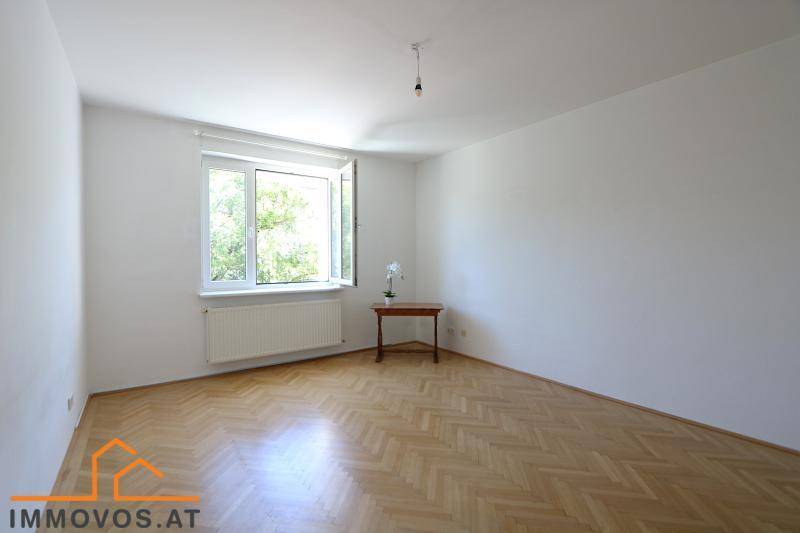 Leeres Zimmer mit Fischgrätparkett, großem Fenster und viel Tageslicht.