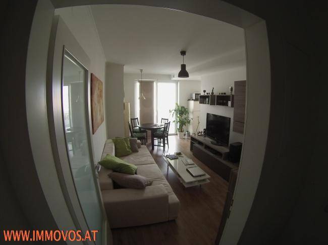 Geräumiges Wohnzimmer mit beigem Sofa, Fernseher und Blick auf den Essbereich und Balkon.