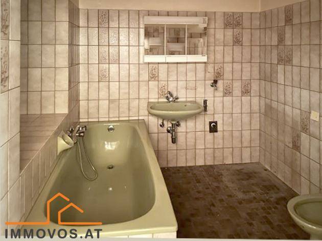 Badezimmer im Originalzustand mit grüner Badewanne, Waschbecken und gemusterten Fliesen.