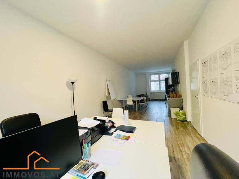 Büroarbeitsplatz mit Schreibtisch und Computer, der sich in einen größeren Raum mit weiteren Arbeitsplätzen öffnet.