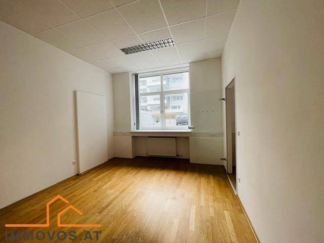 Heller Büroraum mit Holzparkett, weißen Wänden und einem breiten Fenster, das viel Tageslicht hereinlässt.