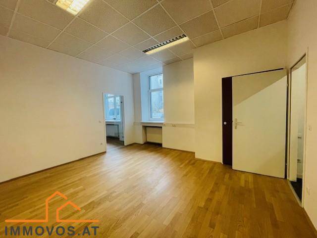 Leeres Büro mit Holzfußboden, weißen Wänden und Zugang zu weiteren Räumen, gut beleuchtet.