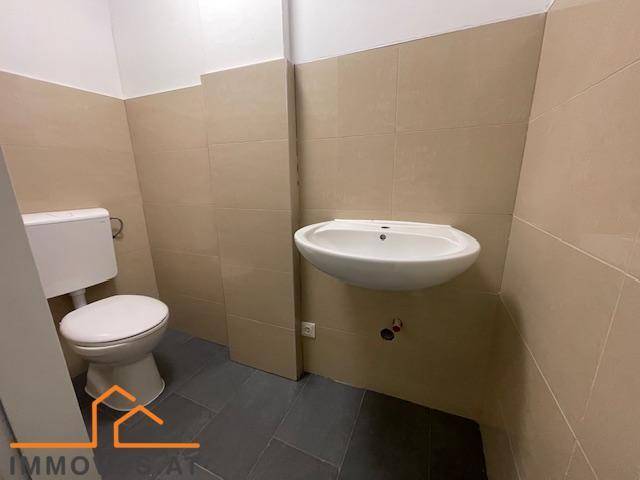 Sauberer Toilettenraum mit WC, wandmontiertem Waschbecken und beigefarbenen Fliesen an Wänden und Boden.