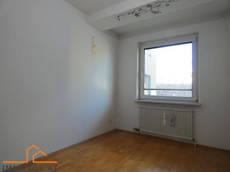 Helles, leeres Zimmer mit Holzboden, weiß gestrichenen Wänden und einem Fenster mit Blick nach draußen.