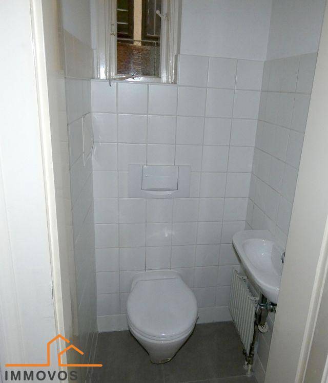 Kleines Gäste-WC mit weißen Wandfliesen, wandhängendem WC und einem kleinen Waschbecken unter einem Fenster.