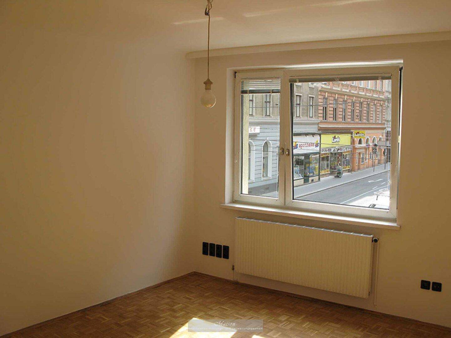 Leeres Zimmer mit Parkettboden und großem Fenster mit Blick auf eine belebte Straße.