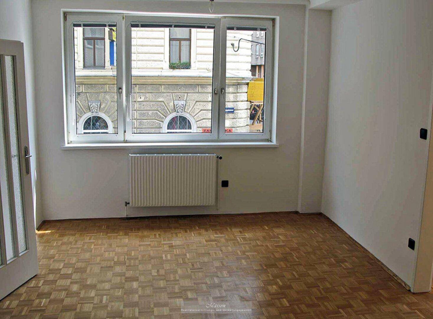 Helles Zimmer mit Parkettboden, Heizkörper und großen Fenstern, die auf die Stadt blicken.