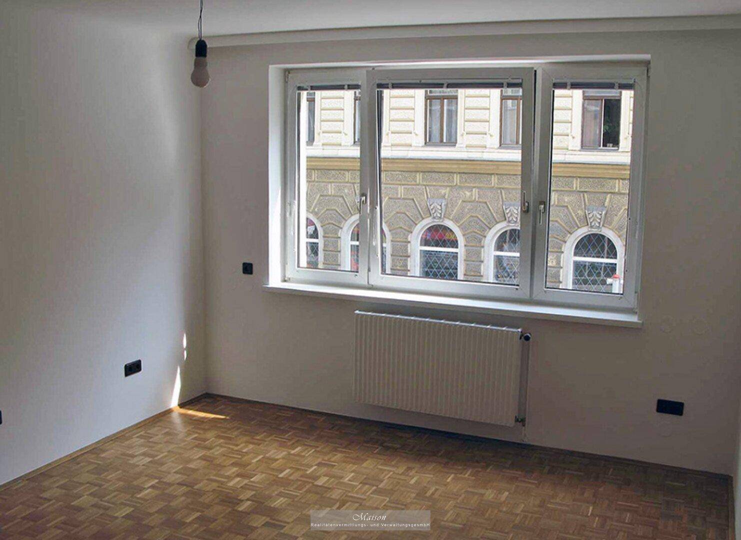 Geräumiges, helles Zimmer mit Parkettboden und breitem Fenster mit Blick auf die gegenüberliegende Fassade.
