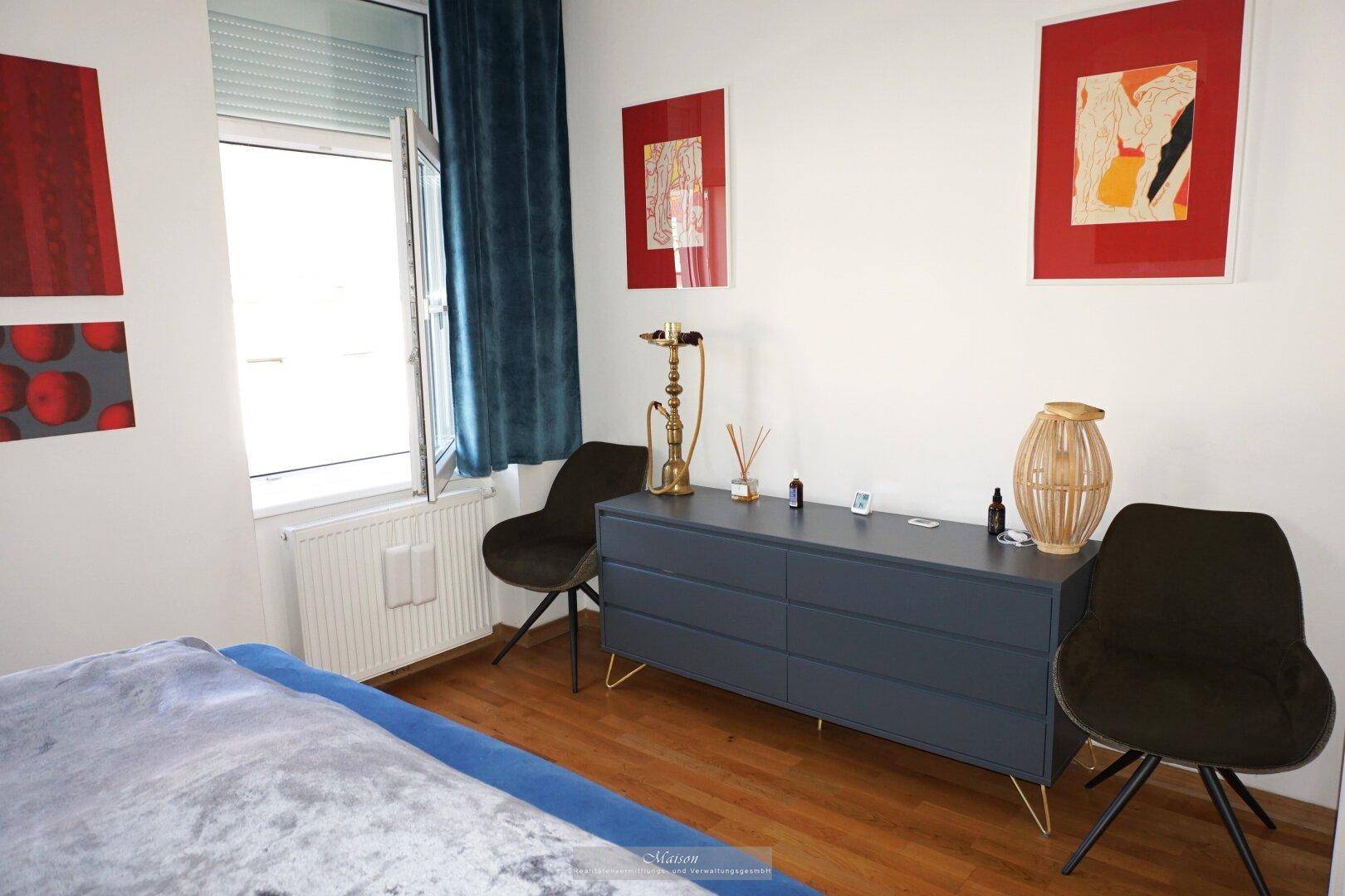 Schlafzimmer mit Kommode, Kunstwerken und einem Fenster, das viel Tageslicht hereinlässt.