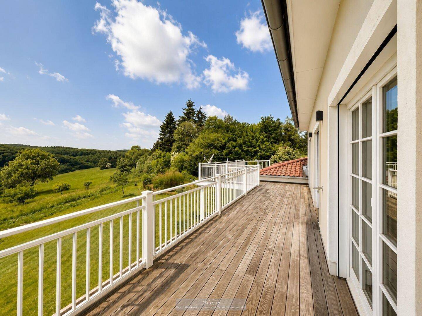 Große Holzterrasse mit weißem Geländer und weitem Blick ins Grüne unter blauem Himmel.