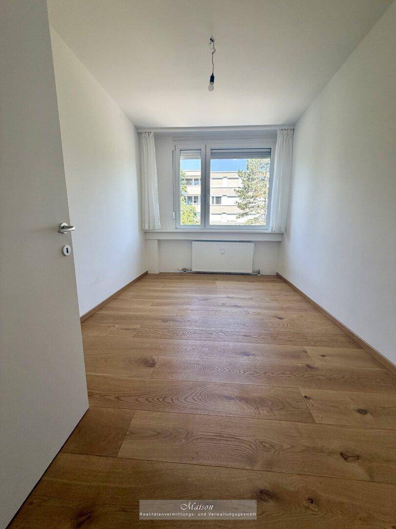 Helles, leeres Zimmer mit Holzparkettboden und einem großen Fenster, das viel Tageslicht hereinlässt.
