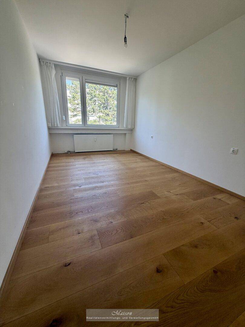 Geräumiges Zimmer mit hellem Holzparkett und großen Fenstern, die einen Blick ins Grüne bieten.