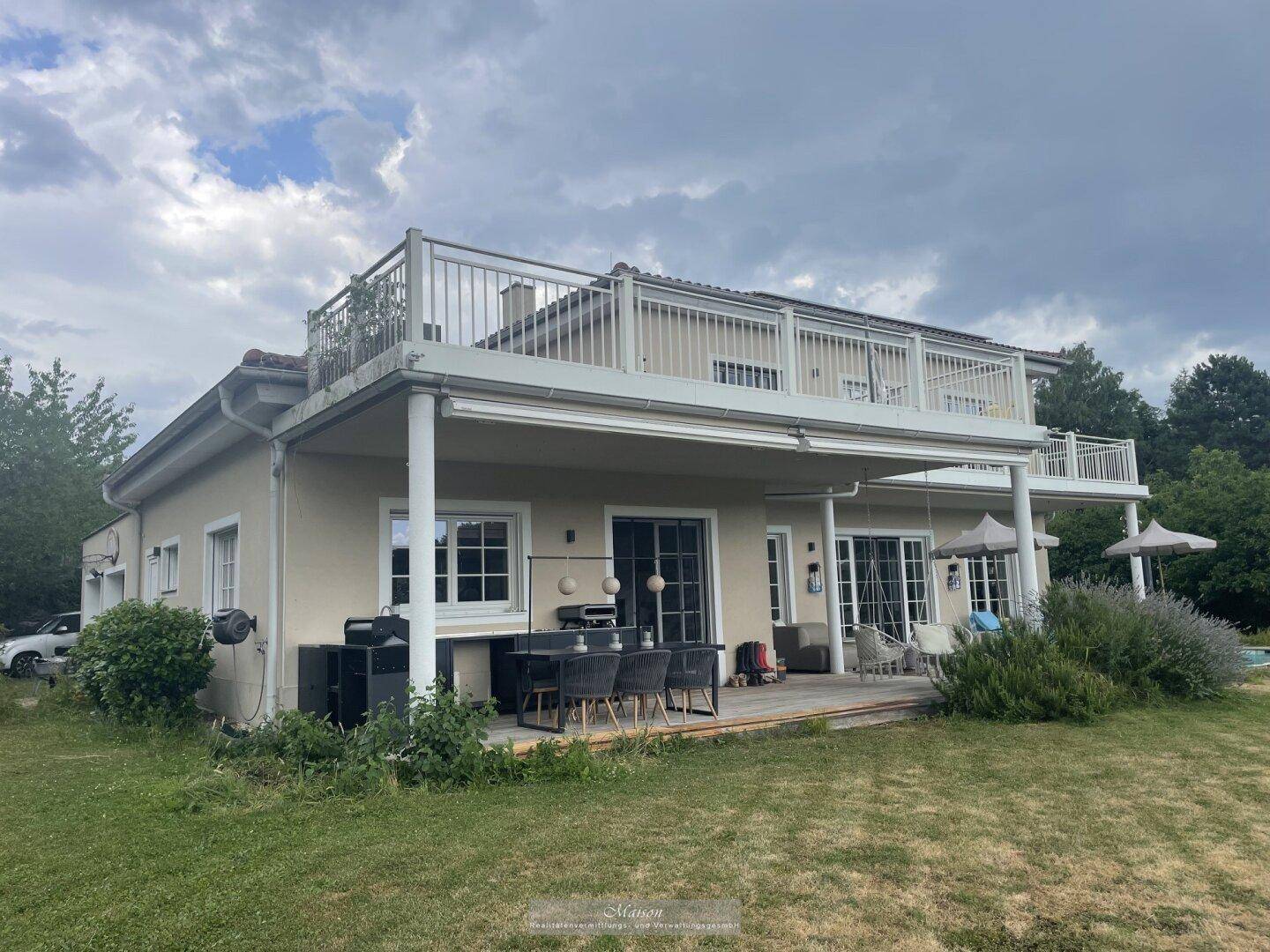 Großzügige Terrasse mit Essbereich und Grill, darüber ein großer Balkon mit weißem Geländer und Blick ins Grüne.