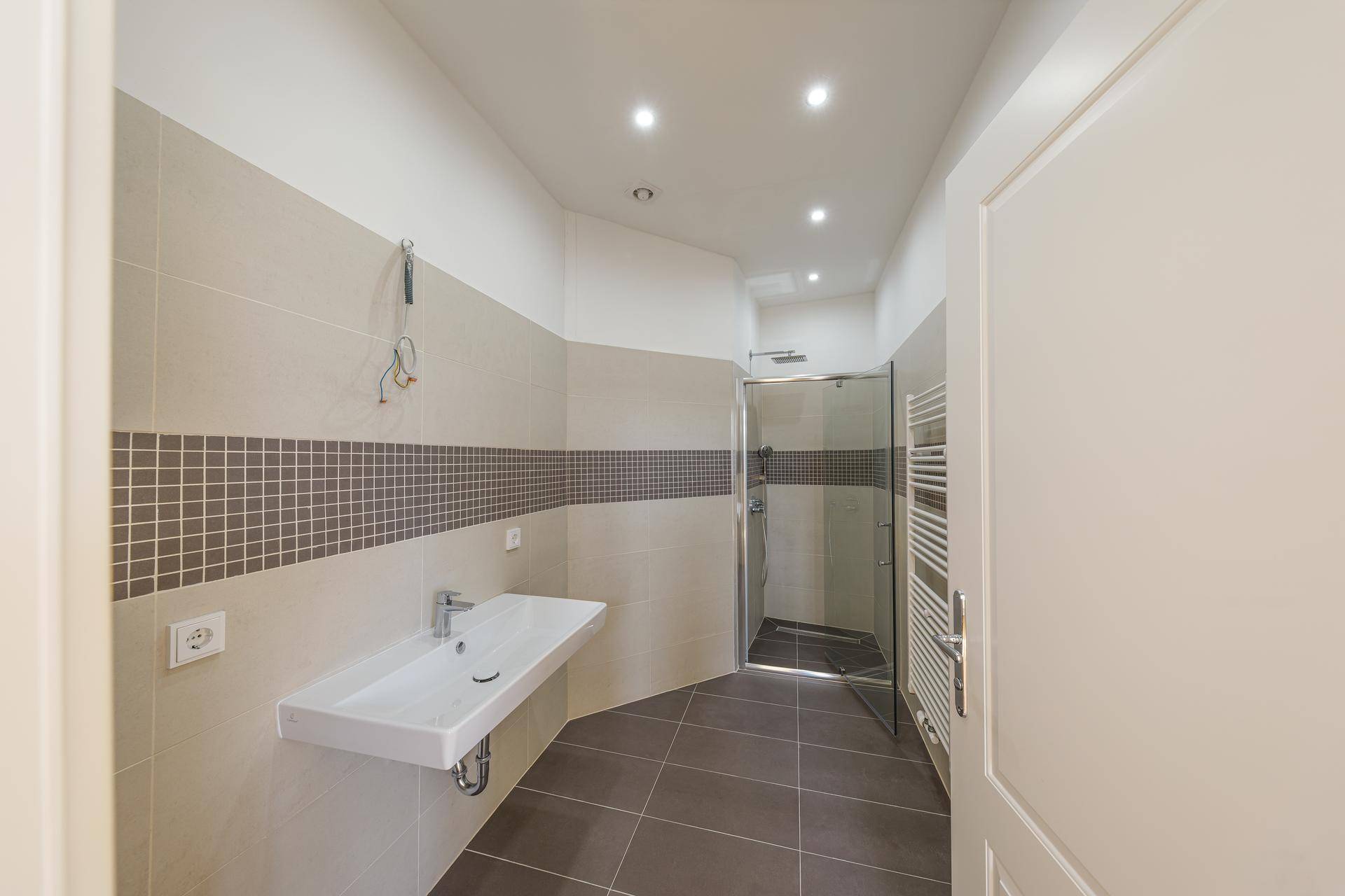 Modernes Badezimmer mit Dusche, wandhängendem Waschbecken und beigen Fliesen.