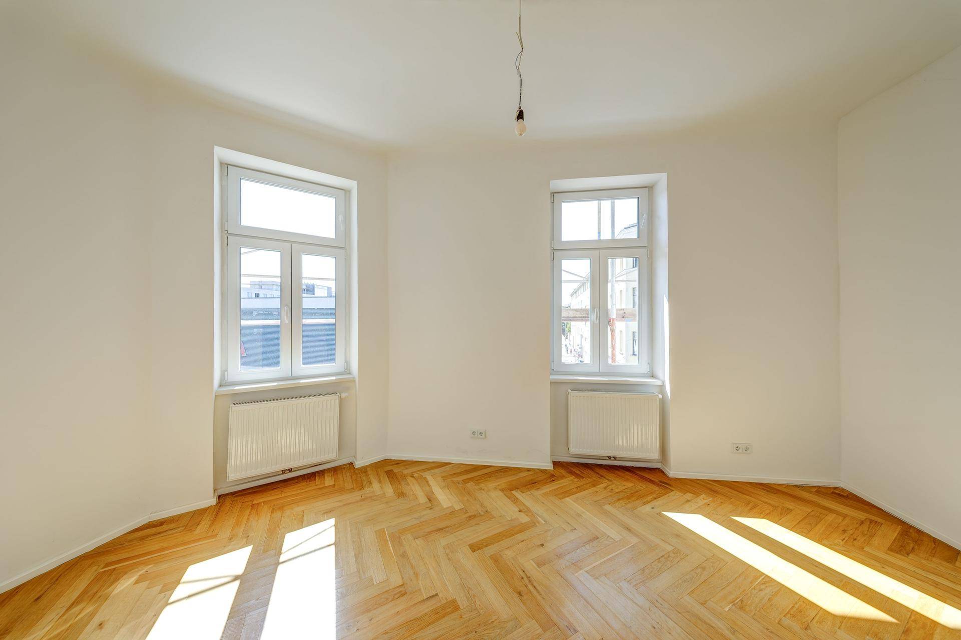 Ruhiges Zimmer mit hellem Fischgrätparkett und zwei Fenstern, ideal als Schlaf- oder Arbeitszimmer.