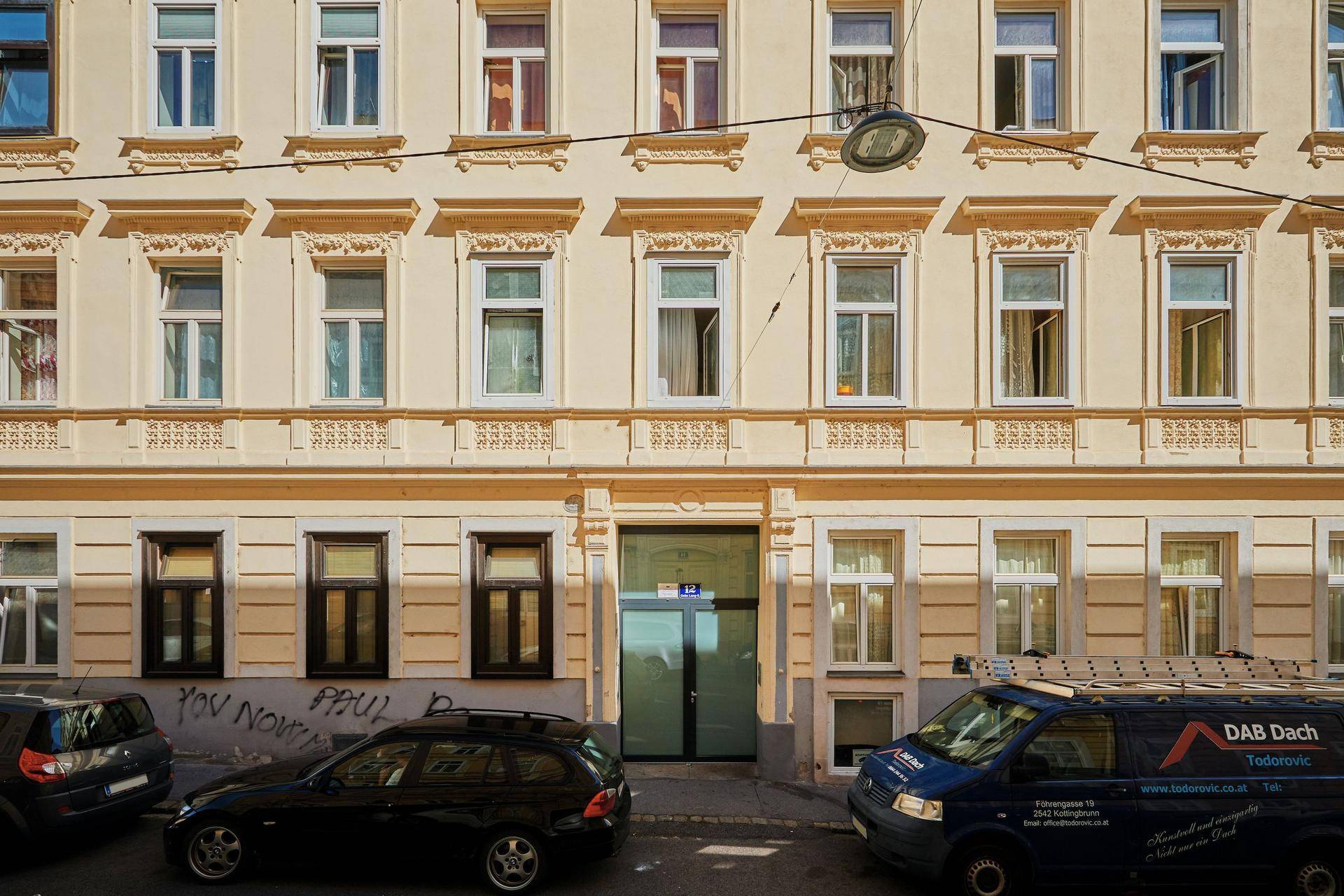 Eingangsbereich des Gebäudes mit großen Fenstern im Erdgeschoss und geparkten Fahrzeugen auf der Straße.