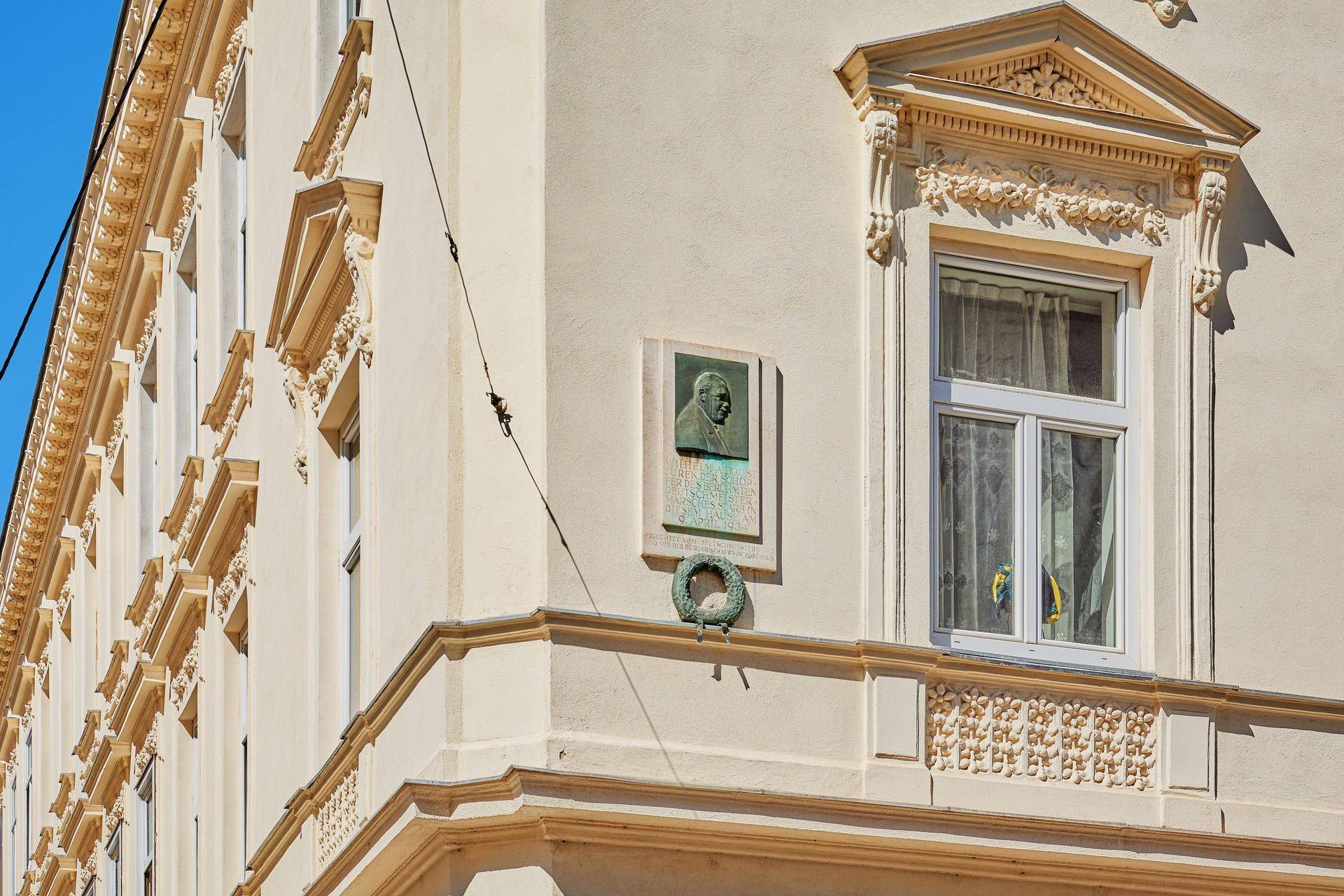 Detailansicht eines Fensters mit kunstvollen Verzierungen und einer Gedenktafel an der Gebäudeecke.