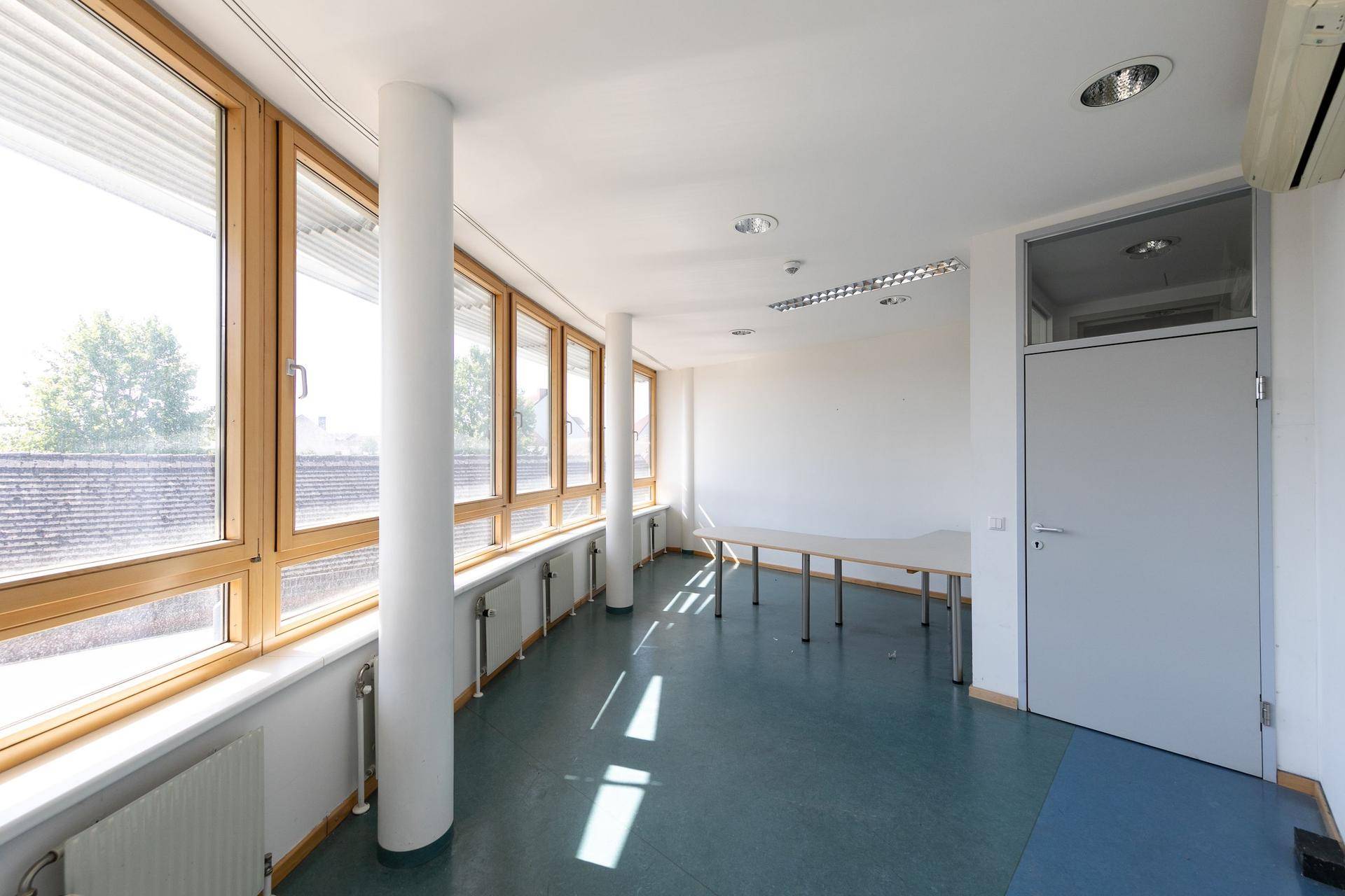 Heller Büroraum mit großen Holzfenstern, blauer Bodenfläche und Klimaanlage.