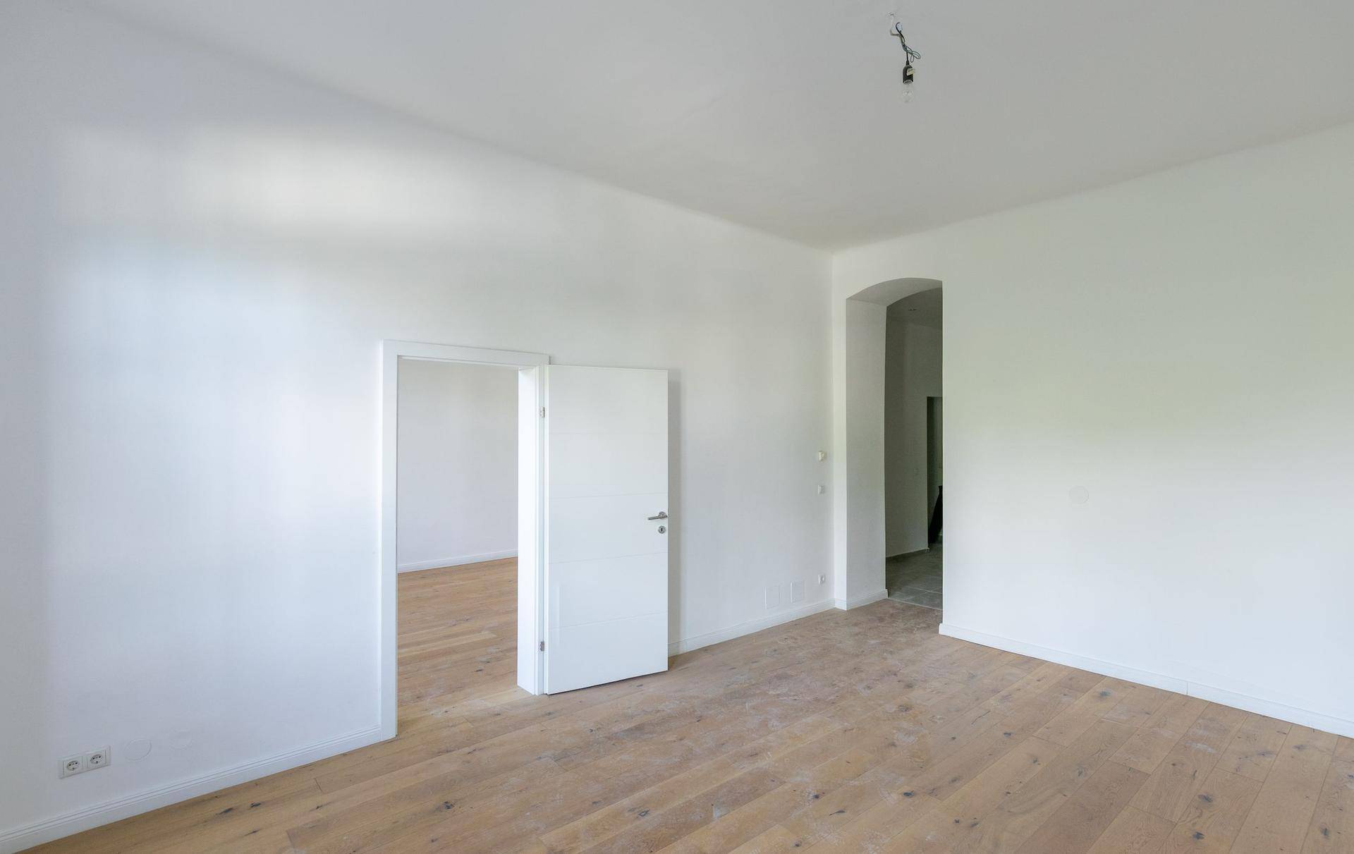 Geräumiges, helles Zimmer mit Holzboden und weiß gestrichenen Wänden, mit Zugang zu weiteren Räumen.
