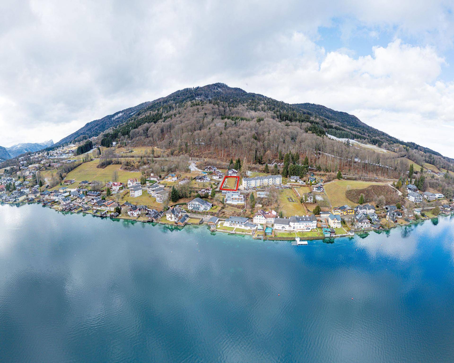 Panoramische Luftaufnahme eines Sees mit umliegenden Bergen und einem Dorf am Ufer.
