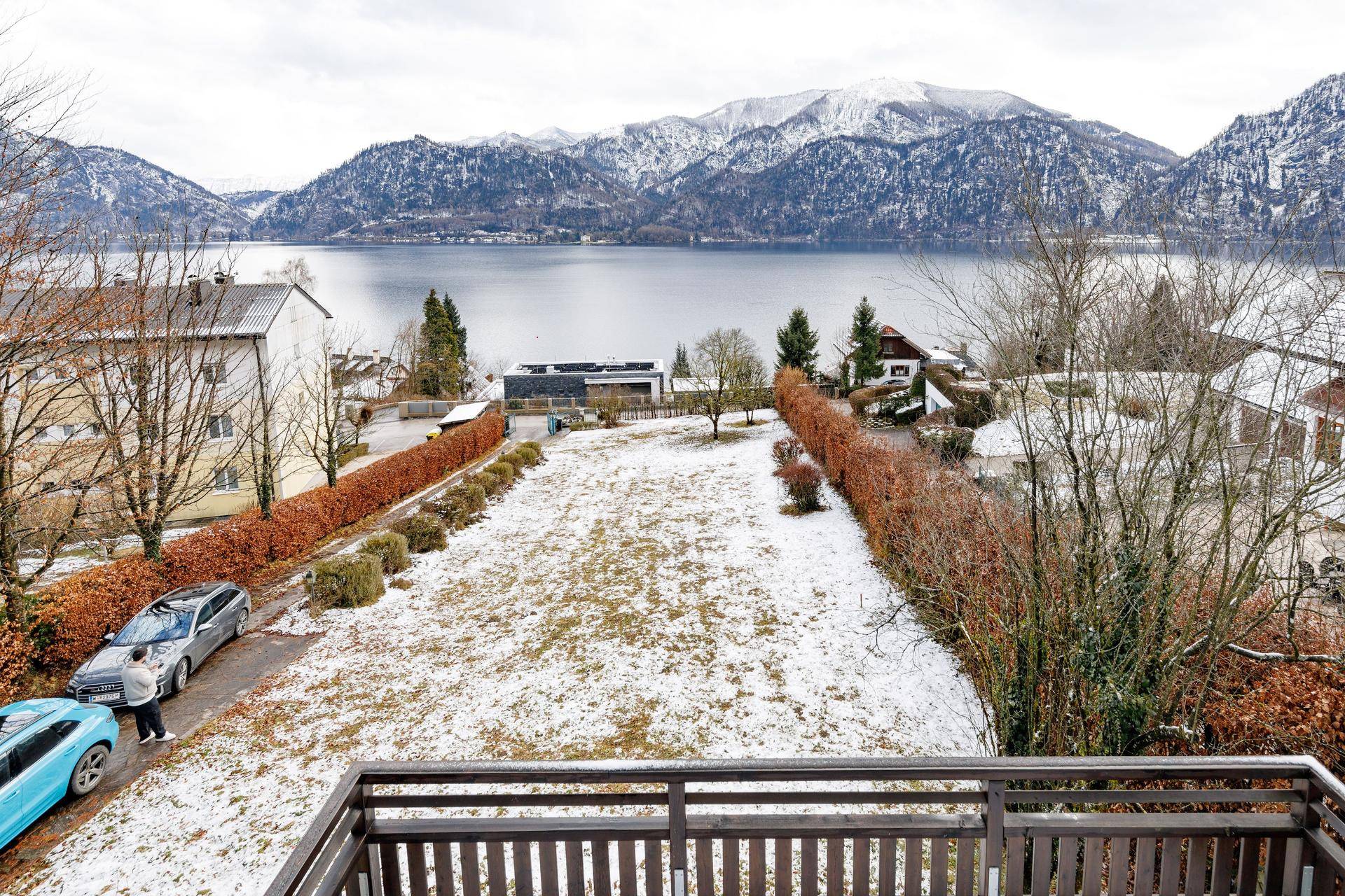 Blick von einem Balkon auf eine verschneite Rasenfläche, den See und die umliegenden Berge.