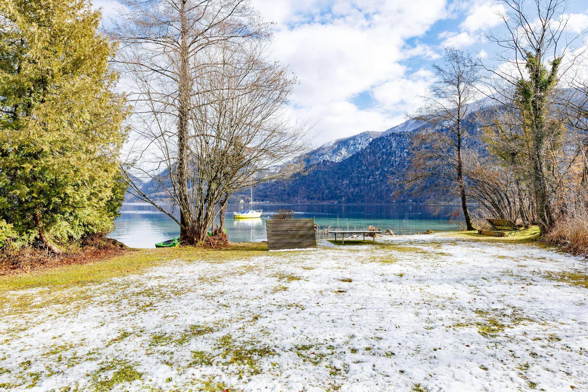 Verschneite Rasenfläche mit Bäumen und einem Picknicktisch, direkt am See mit Bergblick gelegen.