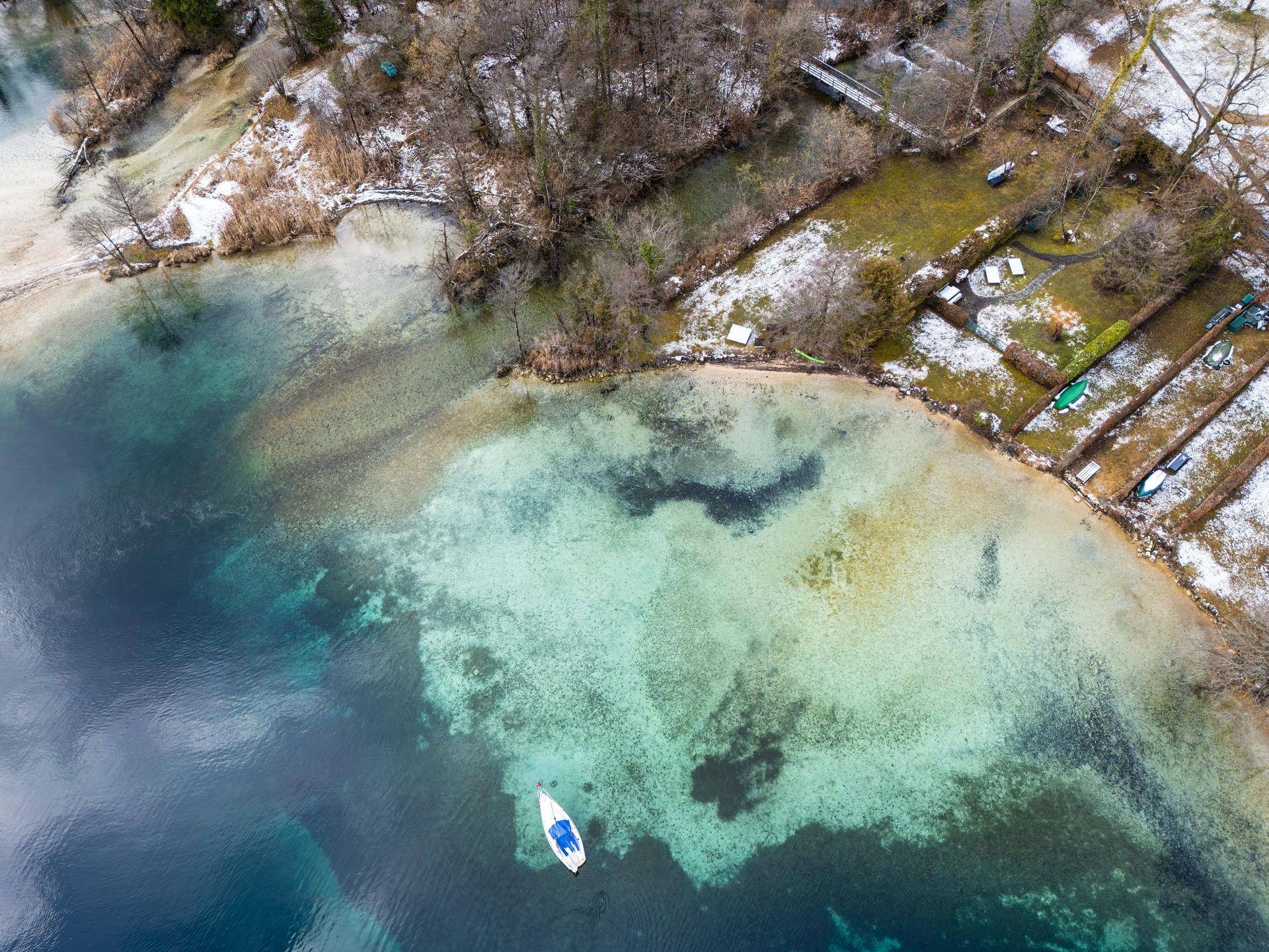 Blick von oben auf das klare, türkisfarbene Wasser des Sees und das schneebedeckte Ufer mit einem Boot.