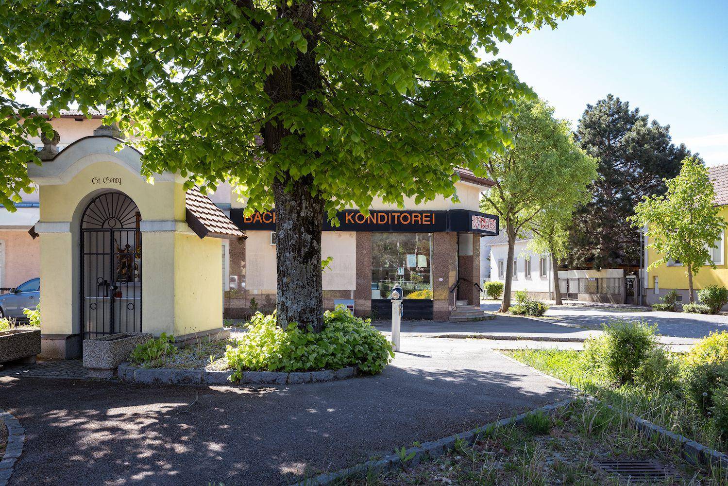 Vorderansicht eines Gebäudes mit Bäckerei, einem großen Baum und einem kleinen Schrein im Vordergrund.