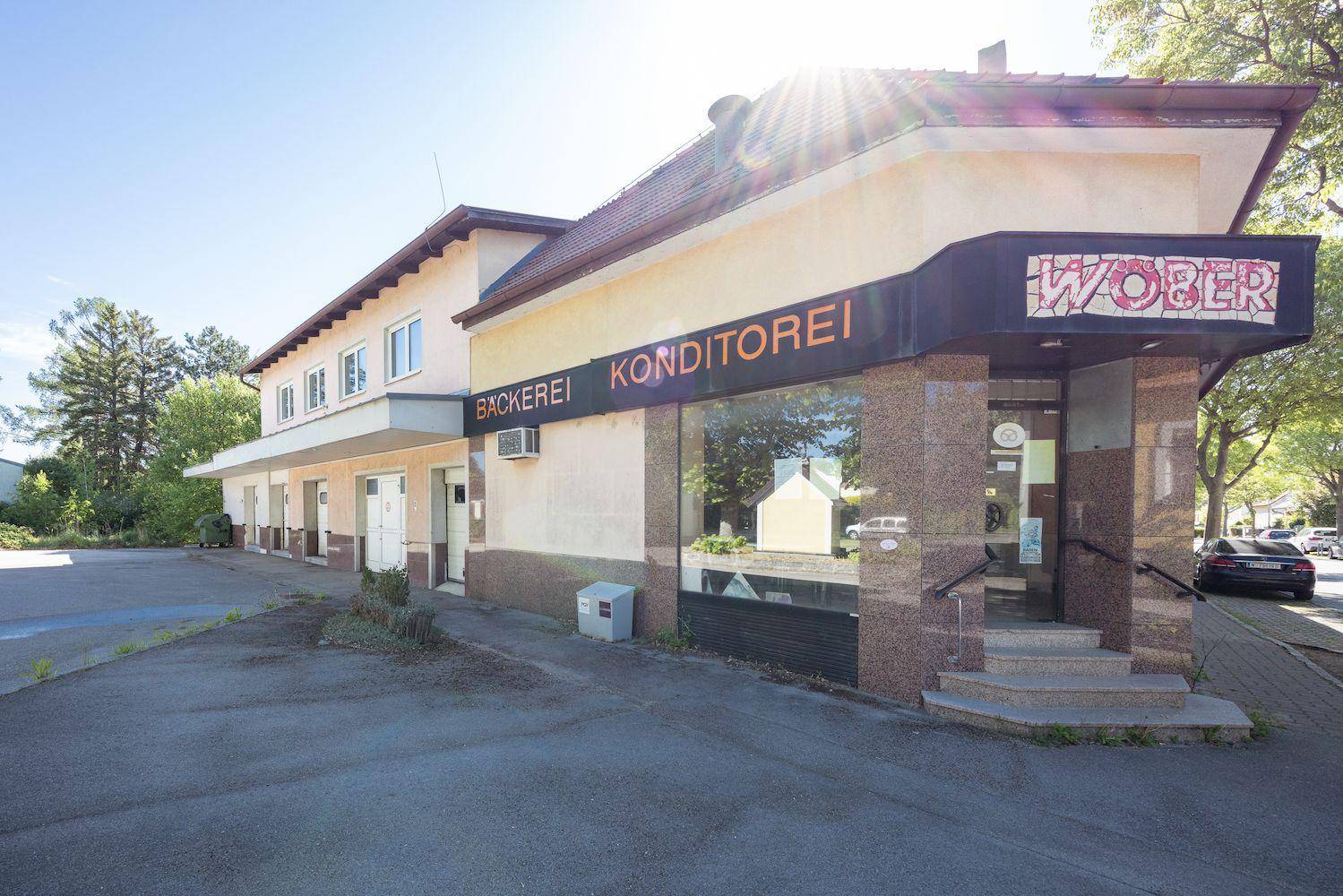 Eckgebäude mit Bäckerei und Konditorei im Erdgeschoss, umgeben von Parkplätzen und Bäumen.