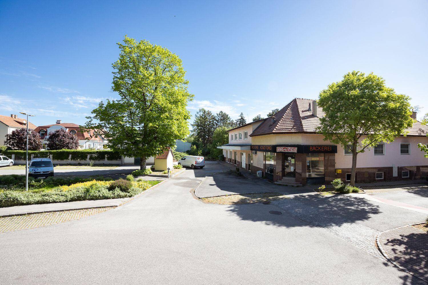 Weitläufige Ansicht eines Gebäudes mit Bäckerei, umgeben von Parkplätzen und viel Grün.