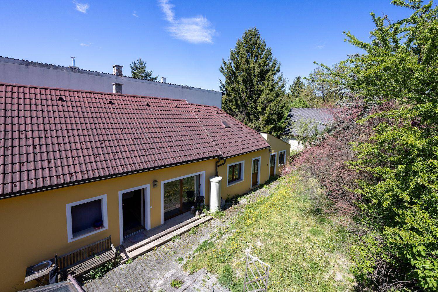 Rückseite eines gelben Hauses mit rotem Ziegeldach und einem verwilderten Gartenbereich.