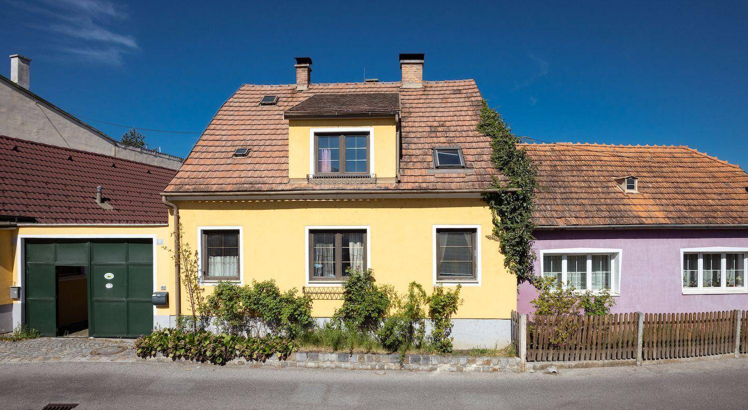 Fassade eines gelben Hauses mit rotem Ziegeldach und einem angrenzenden lila Haus.
