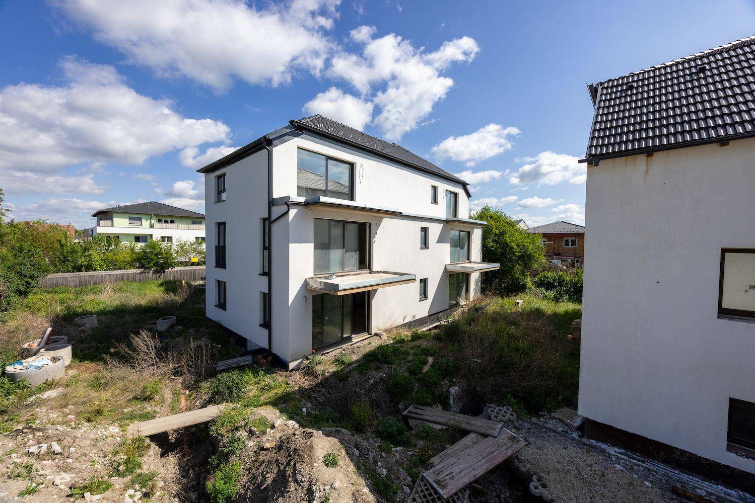 Seitenansicht eines modernen Neubaus mit weißer Fassade und vorbereiteten Balkonen auf einem Hanggrundstück.