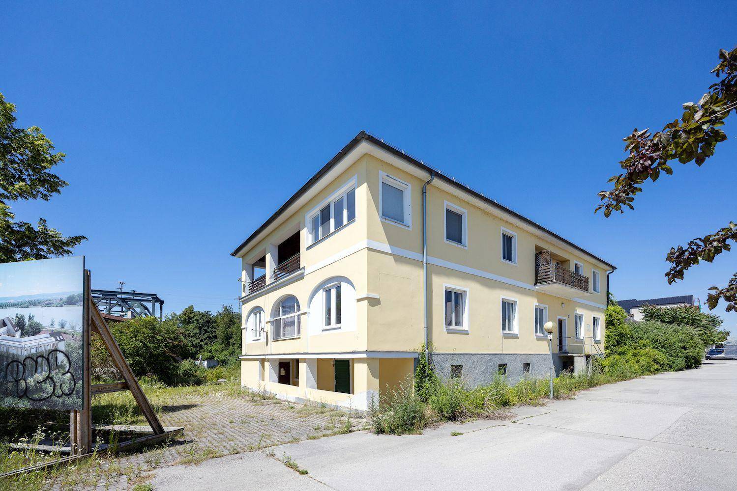 Gelbes Mehrfamilienhaus mit Balkonen und einem Bauschild im Vordergrund unter strahlend blauem Himmel.