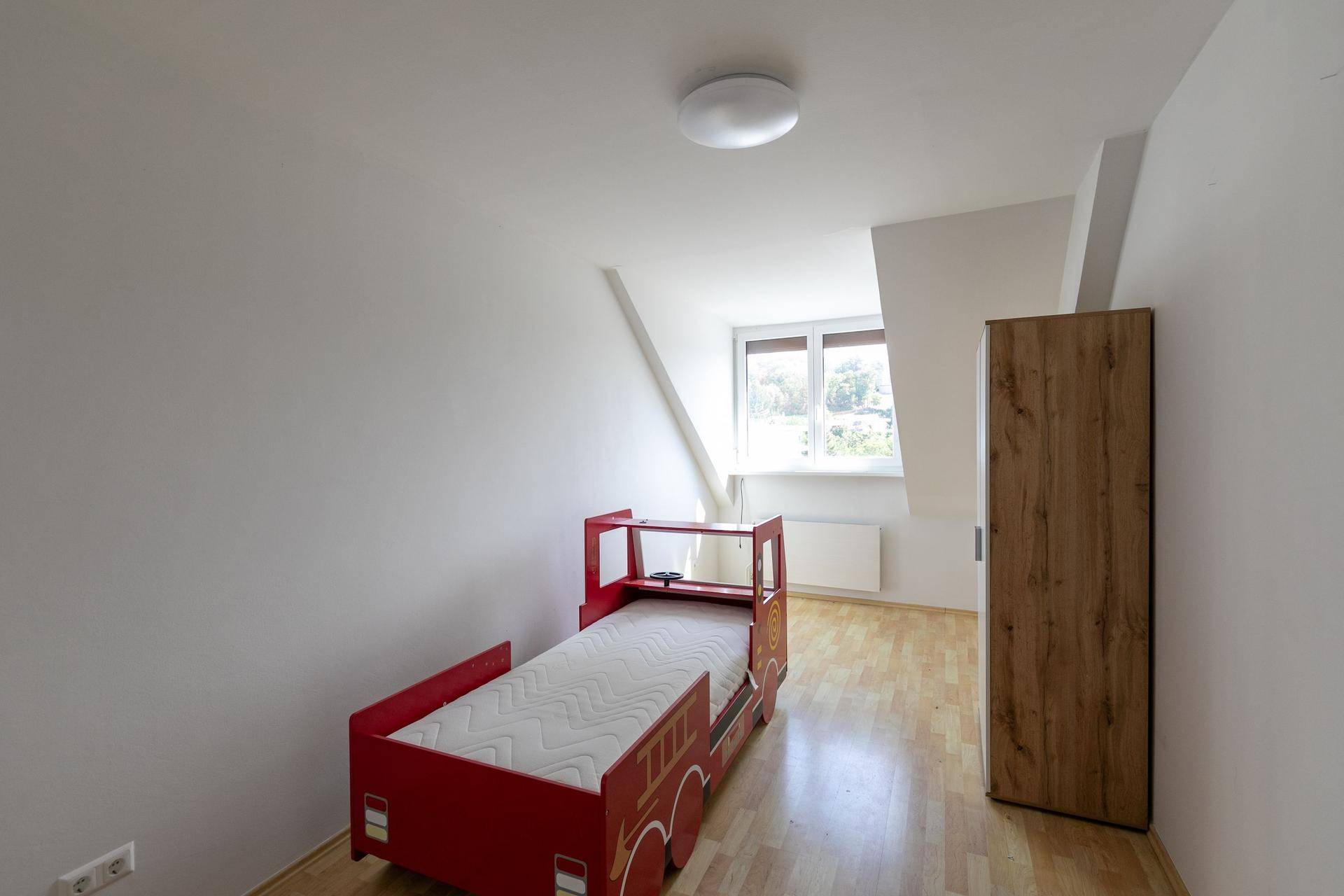 Kinderzimmer mit hellem Parkettboden, einem roten Feuerwehrauto-Bett und Dachschrägen.